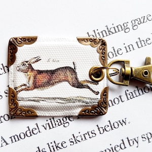 Puede incluir: Llavero rectangular con una ilustración de una liebre corriendo. El llavero tiene un fondo blanco con un patrón texturizado, esquinas decorativas de bronce y un cierre de bronce. La liebre es marrón y beige.