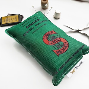 Può includere: Un cuscino per spilli in tessuto verde con un logo Singer Sewing Machines in rosso e bianco. Il cuscino per spilli è pieno di spilli e ha un'etichetta bianca con il testo "Traky Breeks" sul fondo.