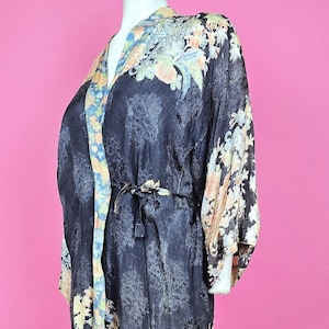 May include: A long, black robe featuring a repeating tonal pattern and a contrasting trim with a floral print in shades of orange, yellow, and blue. The garment includes long sleeves, a V-neckline, and a matching fabric tie belt at the waist.
