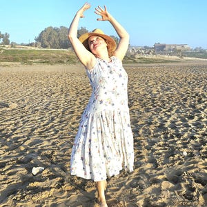 May include: A woman in a white floral dress and a straw hat on a sandy beach, arms raised. The dress features a fitted top and a flowing skirt. The clear blue sky provides a bright backdrop.