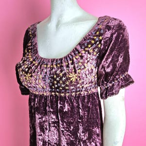 May include: A dark purple velvet dress with short sleeves and a scoop neckline. The bodice features embroidered floral details in gold and yellow. The dress has a gathered waist and a flowing skirt, creating a vintage, bohemian style.