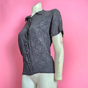 Könnte beinhalten: Ein schwarzes kurzärmeliges Hemd mit floralem Muster. Das Hemd ist auf einer Schaufensterpuppe vor einem rosa Hintergrund ausgestellt. Das Hemd hat einen Kragen und Knöpfe.