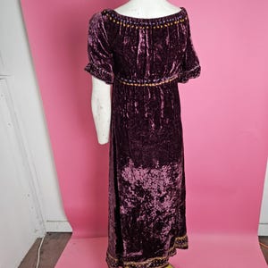 Op de afbeelding: Een lange, donkerpaarse fluwelen jurk met korte mouwen en een ronde hals. De jurk heeft gouden en paarse geborduurde details rond de halslijn, taille en zoom. De jurk wordt op een mannequin getoond.