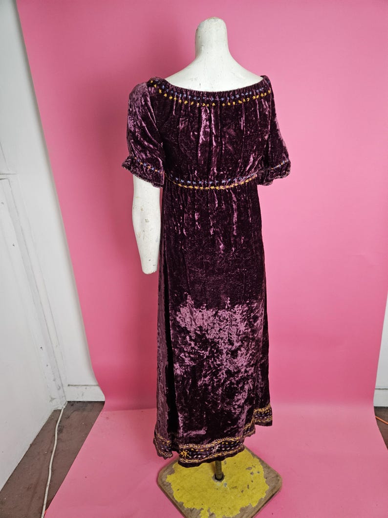 Op de afbeelding: Een lange, donkerpaarse fluwelen jurk met korte mouwen en een ronde hals. De jurk heeft gouden en paarse geborduurde details rond de halslijn, taille en zoom. De jurk wordt op een mannequin getoond.