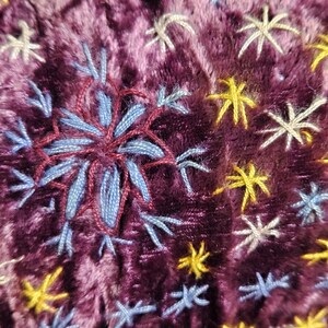 Op de afbeelding: Close-up van een pluche, diep paarse stof met geborduurde sneeuwvlok- en sterrenpatronen. De centrale sneeuwvlok is een gedetailleerde combinatie van blauw en bordeauxrood garen, omringd door kleinere gele, witte en blauwe sterren.