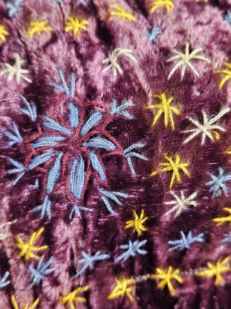 Op de afbeelding: Close-up van een pluche, diep paarse stof met geborduurde sneeuwvlok- en sterrenpatronen. De centrale sneeuwvlok is een gedetailleerde combinatie van blauw en bordeauxrood garen, omringd door kleinere gele, witte en blauwe sterren.