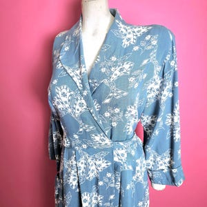 Könnte beinhalten: Hellblaues Kleid mit weißem Blumenmuster. Das Kleid hat lange Ärmel, einen Wickel-Oberteil und einen plissierten Rock. Der Stoff wirkt weich und fließend. Das Kleid wird auf einer Schaufensterpuppe präsentiert.