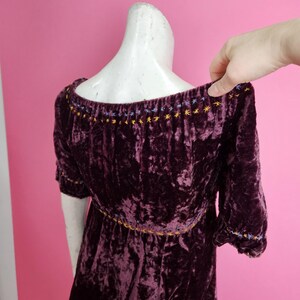 Op de afbeelding: Een donkerpaarse fluwelen jurk met korte mouwen. De halslijn en mouwen zijn versierd met geborduurde ster- en stippatronen in geel, blauw en paars. De jurk wordt gepresenteerd op een mannequin tegen een roze achtergrond.