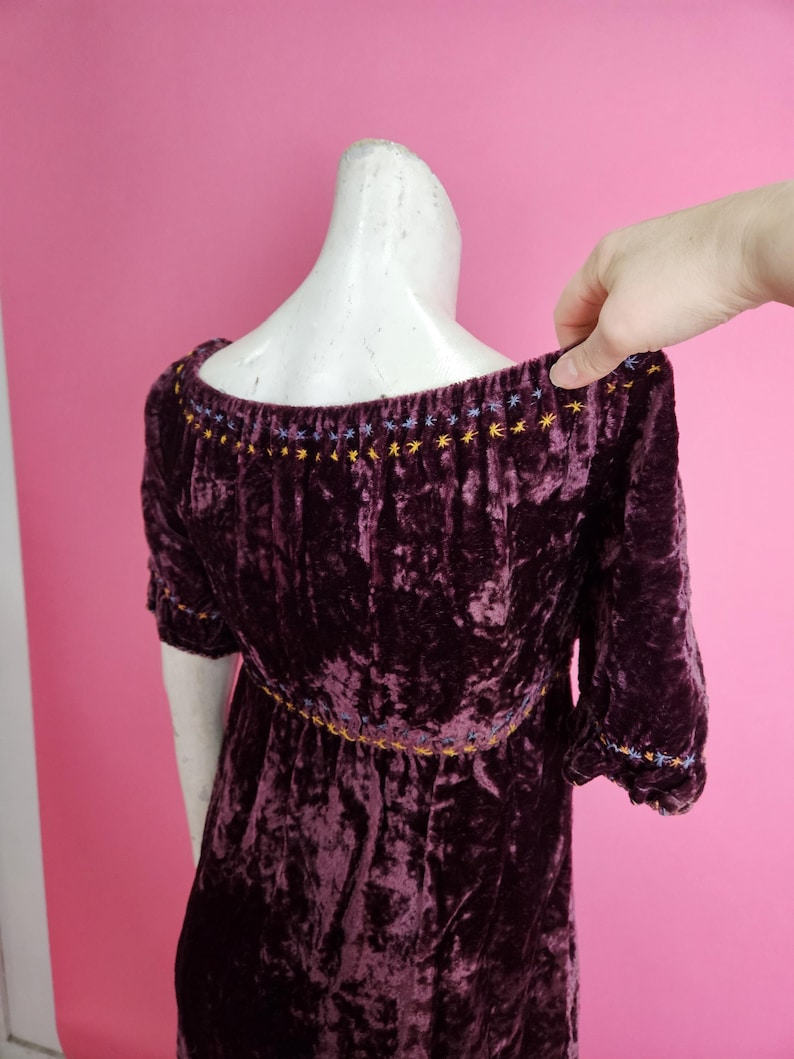 Op de afbeelding: Een donkerpaarse fluwelen jurk met korte mouwen. De halslijn en mouwen zijn versierd met geborduurde ster- en stippatronen in geel, blauw en paars. De jurk wordt gepresenteerd op een mannequin tegen een roze achtergrond.