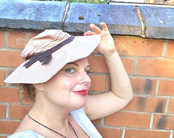 Puede incluir: Un sombrero de sol de color beige claro con una cinta y un lazo de color marrón oscuro. El sombrero presenta una copa fruncida y un ala ancha ligeramente inclinada hacia abajo. También se ven un collar de cordón oscuro con un colgante verde.