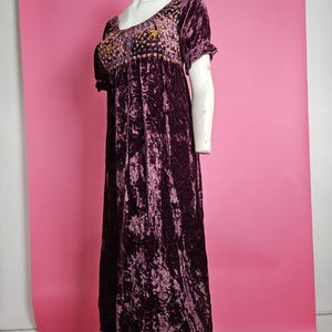 Op de afbeelding: Een lange, donkerpaarse fluwelen jurk met korte mouwen. De bodice en mouwen hebben gedetailleerde gouden borduursels. De jurk wordt getoond op een mannequin tegen een roze achtergrond, wat het vintage ontwerp benadrukt.