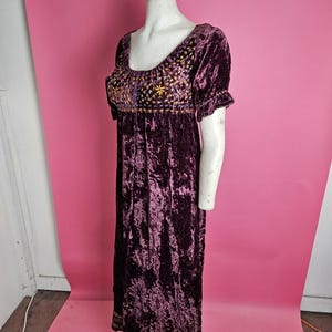 Op de afbeelding: Een donkerpaarse fluwelen jurk met korte mouwen en een ronde halslijn. De lijfjes en zoom zijn versierd met gouden borduursels. De jurk wordt gepresenteerd op een mannequin tegen een roze achtergrond.