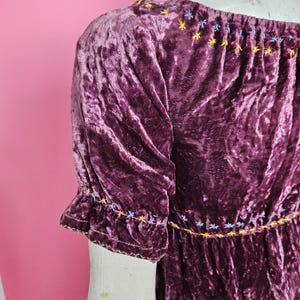 Op de afbeelding: Een pruimkleurige fluwelen jurk met korte mouwen. De jurk heeft geborduurde stervormige details in geel, blauw en wit langs de halslijn en manchetten. De jurk heeft een geplooid taille en een zachte, getextureerde uitstraling.