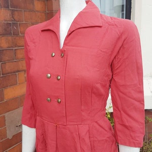 Puede incluir: Vestido vintage color coral con cuello y mangas tres cuartos. El vestido presenta un diseño cruzado con botones dorados y una falda plisada. La tela tiene un sutil patrón texturizado. El vestido se exhibe en un maniquí.