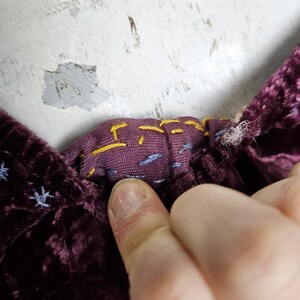 Op de afbeelding: Close-up van een diep paarse fluwelen stof met een contrasterende patroonvoering. De voering heeft gele en blauwe stikseldetails. De stof wordt vastgehouden door de vinger van een persoon.