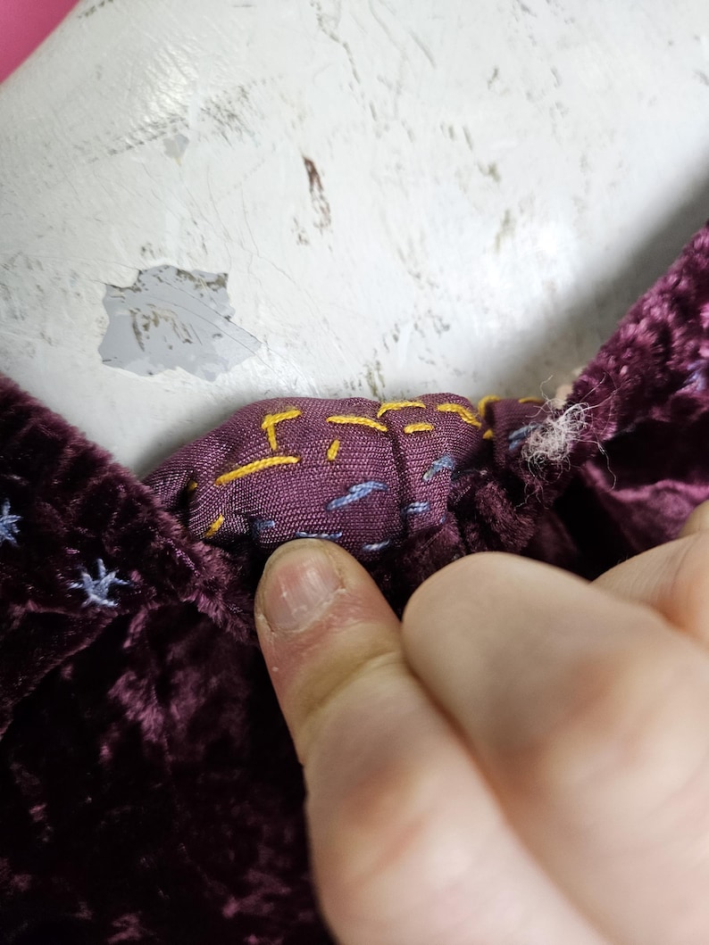 Op de afbeelding: Close-up van een diep paarse fluwelen stof met een contrasterende patroonvoering. De voering heeft gele en blauwe stikseldetails. De stof wordt vastgehouden door de vinger van een persoon.