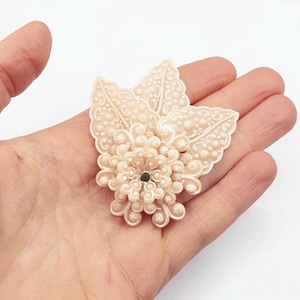 Può includere: Un delicato spilla floreale di colore avorio con un design testurizzato in rilievo. La spilla presenta tre foglie sovrapposte con un fiore centrale composto da piccoli petali arrotondati.