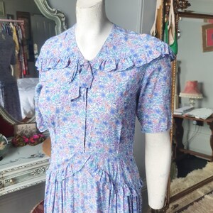 Op de afbeelding: Een vintage jurk met een bloemenprint in tinten blauw, roze en wit. De jurk heeft een geruite kraag en korte mouwen. Het is een midi-jurk met een getailleerd lijfje en een volle rok.