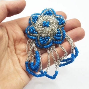 Op de afbeelding: Een blauwe en witte kralenbloem met een franje van kralen. De bloem is gemaakt van kleine, ronde kralen die aan elkaar zijn genaaid om een ​​driedimensionaal ontwerp te creëren.