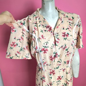 Op de afbeelding: Een vintage perzik kleurige jurk met een bloemenpatroon van roze en witte bloemen en groene bladeren. De jurk heeft korte mouwen, een kraag en witte afwerking. De jurk wordt op een mannequin getoond.