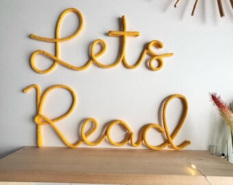 Puede incluir: Un letrero decorativo de pared hecho de cordón tejido amarillo que forma el texto cursivo "Let's Read". Las letras están dispuestas en dos líneas, con "Let's" arriba y "Read" abajo, presentando una apariencia texturizada similar a una cuerda.