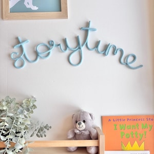 May include: A light blue "storytime" wall decoration made of rope. Below, a wooden shelf holds a gray teddy bear and children's books, including "I Want My Potty!" and "The Very Hungry Caterpillar."