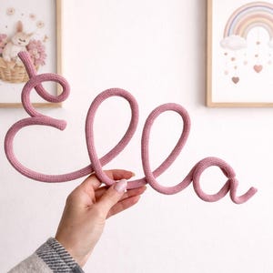 Puede incluir: Un cordón tejido de color rosa viejo que forma el nombre Ella en letra cursiva. Las letras están hechas de una sola pieza continua de alambre cubierto de tela, con una superficie tejida y texturizada que crea un aspecto suave y redondeado.