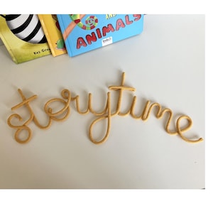 Pode incluir: Um letreiro de parede amarelo-mostarda feito de um comprimento contínuo de cordão de malha, soletrando a palavra storytime em letra cursiva minúscula. As letras estão conectadas em uma única linha fluida, criando um enfeite de parede.