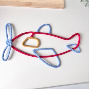 Op de afbeelding: Een wanddecoratie in de vorm van een vliegtuig gemaakt van rood, lichtblauw en mosterdgeel touw. Het rode touw vormt de romp, terwijl lichtblauw touw de propeller, vleugels en staart vormt. Een mosterdgeel touw vormt een raam in het midden.