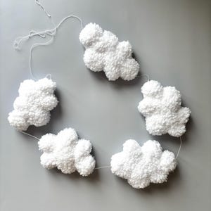 May include: A white cloud garland made from a fluffy, textured material. The garland features five cloud shapes strung together with white thread, ideal for nursery or party decor. The clouds are arranged in a circular pattern against a grey background.