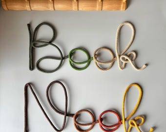 Puede incluir: Dos obras de arte con palabras de cuerda que deletrean "Book Nook" en verde, beige, marrón y amarillo. Un perchero de madera con cinco clavijas está encima de las palabras. Hojas de eucalipto en la parte superior de la imagen.