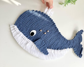 Puede incluir: Un adorno de pared de ballena en macramé azul con una parte inferior de flecos blancos, un ojo negro y blanco y pequeñas cuentas de madera en el costado. La ballena está hecha de cuerda gruesa con una apariencia texturizada y acanalada.