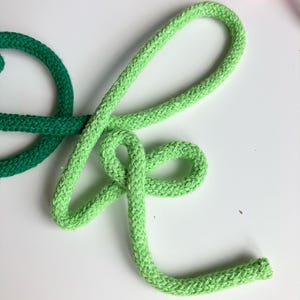 Puede incluir: Una cuerda trenzada de color verde claro con forma de letra K cursiva. La cuerda tiene una superficie tejida texturizada y est&aacute; colocada junto a un segmento de cuerda verde oscuro a la izquierda y una peque&ntilde;a parte de un segmento rosa arriba.