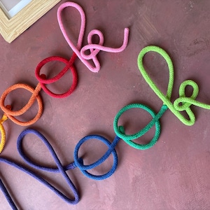 Puede incluir: Un letrero decorativo de pared hecho de cuerda trenzada de colores, que deletrea la palabra "book" en rosa, rojo y naranja, y la palabra "nook" en verde, azul verdoso y azul. Las letras est&aacute;n formadas en un estilo de escritura cursiva.