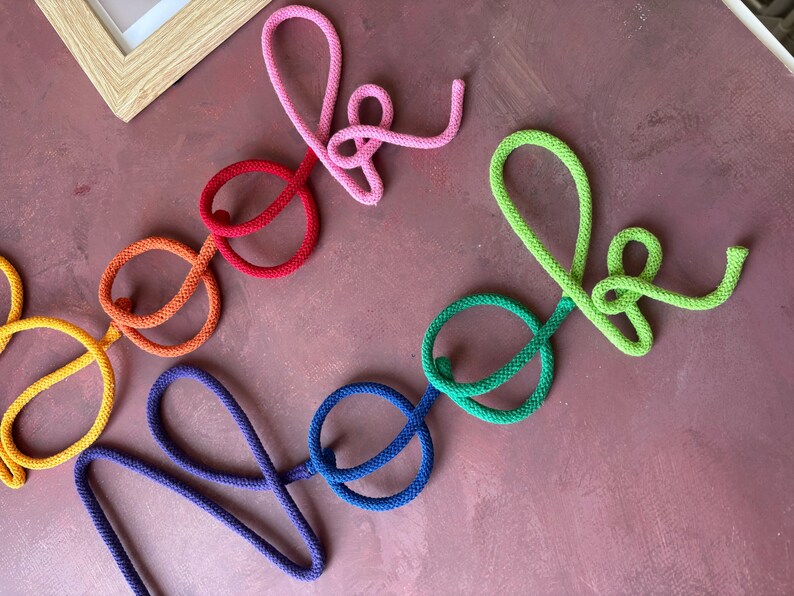 Puede incluir: Un letrero decorativo de pared hecho de cuerda trenzada de colores, que deletrea la palabra "book" en rosa, rojo y naranja, y la palabra "nook" en verde, azul verdoso y azul. Las letras est&aacute;n formadas en un estilo de escritura cursiva.