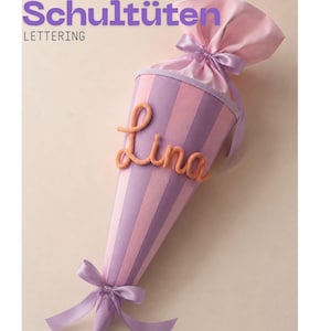 Könnte beinhalten: Eine kegelförmige Schultüte mit vertikalen Streifen in abwechselndem Helllila und Rosa. Der obere Teil ist mit einem passenden lila Satinband gerafft. Der Name Lina ist in orangefarbener Schreibschrift angebracht. Text: Schultüten LETTERING.
