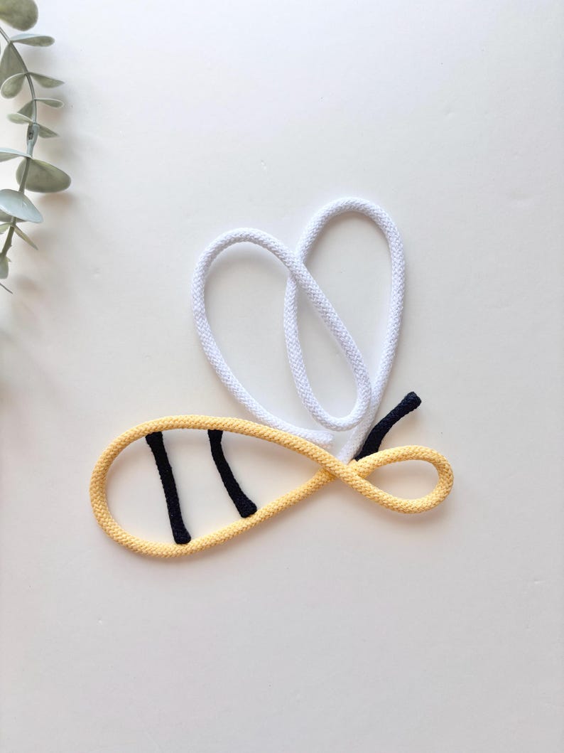 May include: A whimsical bee-shaped rope design. The bee's body is yellow with two black stripes, and the wings are white. The bee's stinger is black. The design is set against a white background.