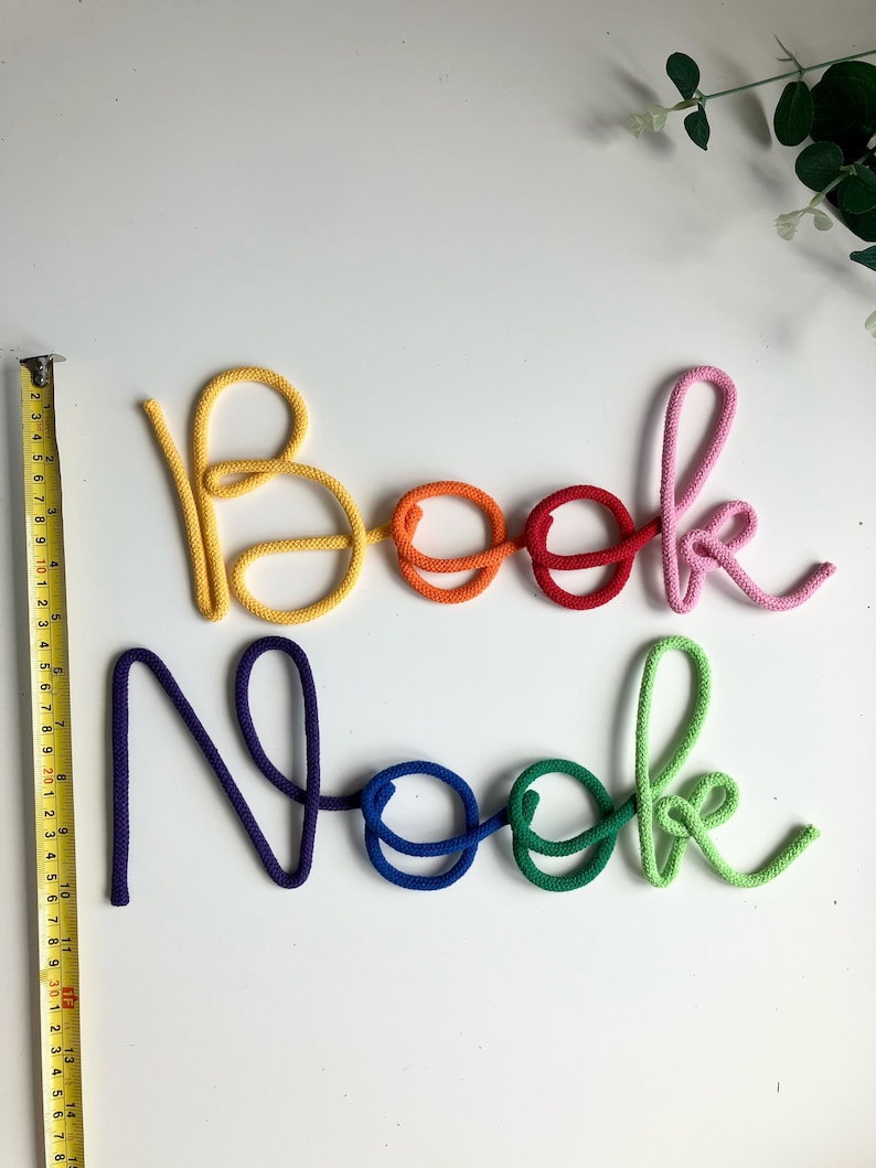 Puede incluir: Decoraci&oacute;n de pared que deletrea las palabras Book Nook en letra cursiva hecha de cuerda colorida. Las letras son amarillas, naranjas, rojas, rosas, moradas, azules y verdes. Una cinta m&eacute;trica amarilla est&aacute; colocada verticalmente junto a las letras.