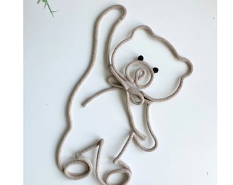 May include: A wall-mounted bear silhouette crafted from a single continuous length of beige rope. The design features a rounded head, a small bow at the neck, and two small black circular eyes. The rope forms the outline of the bear's body.