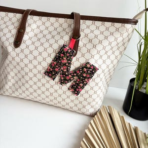 Puede incluir: Un bolso tote beige con un patrón geométrico marrón y asas de cuero marrón. Una etiqueta con estampado floral con una correa rosa cuelga del asa. La etiqueta tiene un fondo negro con flores rosas y rojas.