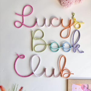 May include: A wall decoration spelling out "Lucy's Book Club" in colorful, rope-like letters. The letters are in shades of pink, orange, yellow, green, blue, and purple. A cluster of pink pom-poms is in the upper right corner.