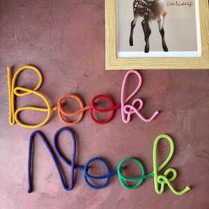 Puede incluir: Un letrero de pared que forma las palabras Book Nook en letra cursiva. Las letras est&aacute;n hechas de cuerda colorida en amarillo, naranja, rojo, rosa, morado, azul, verde y verde lima. Arriba hay un cuadro de un cervatillo con el texto Be curious.
