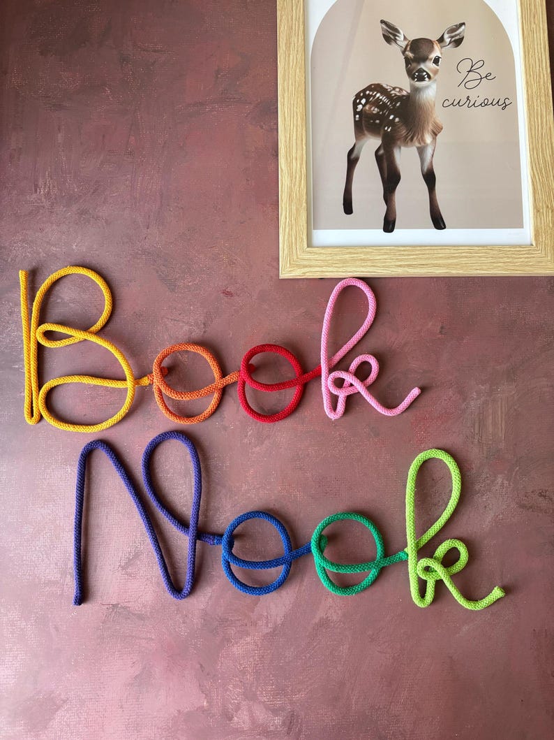 Puede incluir: Un letrero de pared que forma las palabras Book Nook en letra cursiva. Las letras est&aacute;n hechas de cuerda colorida en amarillo, naranja, rojo, rosa, morado, azul, verde y verde lima. Arriba hay un cuadro de un cervatillo con el texto Be curious.