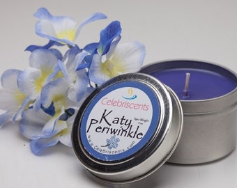 May include: A small tin candle with a purple wax and a silver lid. The lid has a label that says "Celebriscents Katy Periwinkle" with a website address. The candle is next to a bouquet of blue and white flowers.