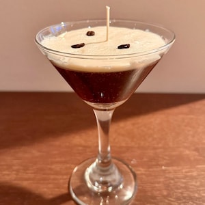 May include: A clear glass martini glass filled with a brown and white layered candle. The candle has a single wick and is decorated with coffee beans.