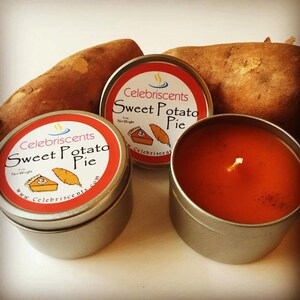 May include: Two silver tins of candles with orange wax and a single wick. The labels on the tins read "Celebriscents Sweet Potato Pie" with a small illustration of a sweet potato pie. Two whole sweet potatoes are in the background.