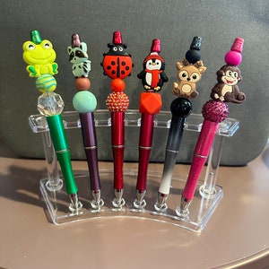 Può includere: Un set di sei penne colorate con diversi animali come topper. Le penne sono esposte in un supporto di plastica trasparente. I topper includono una rana, un procione, una coccinella, un pinguino, un cervo e una scimmia.