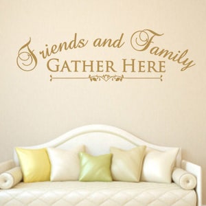 May include: A cream-colored sofa with a tufted seat and rolled armrests is adorned with five throw pillows in shades of yellow and green. The wall above the sofa features the text "Friends and Family Gather Here" in a decorative font.