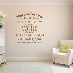 Op de afbeelding: Muursticker met een citaat uit Mattheüs 4:4, in bruine letters. Het citaat luidt: 'De mens zal niet van brood alleen leven, maar van elk woord dat uit de mond van God komt.' Een witte boekenkast en een fauteuil zijn ook zichtbaar.