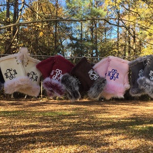 May include: Eight pairs of fleece fingerless gloves with faux fur trim, hanging on a clothesline. Each pair is a different color and has a monogram embroidered on the cuff. The colors are black, white, burgundy, brown, pink, gray, and blue.